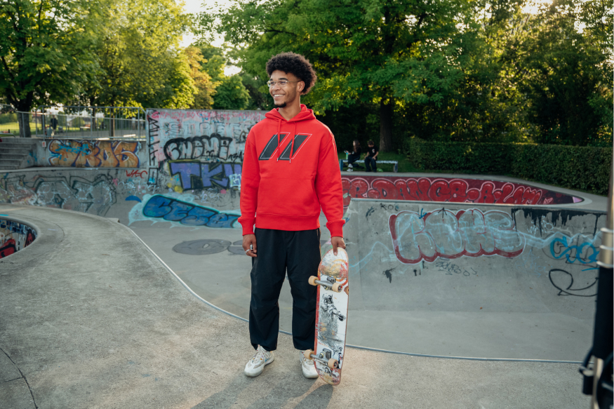 Sportstadt hoodie a red worn by a young man standing in the skate park. He holds a skateboard in his left hand.